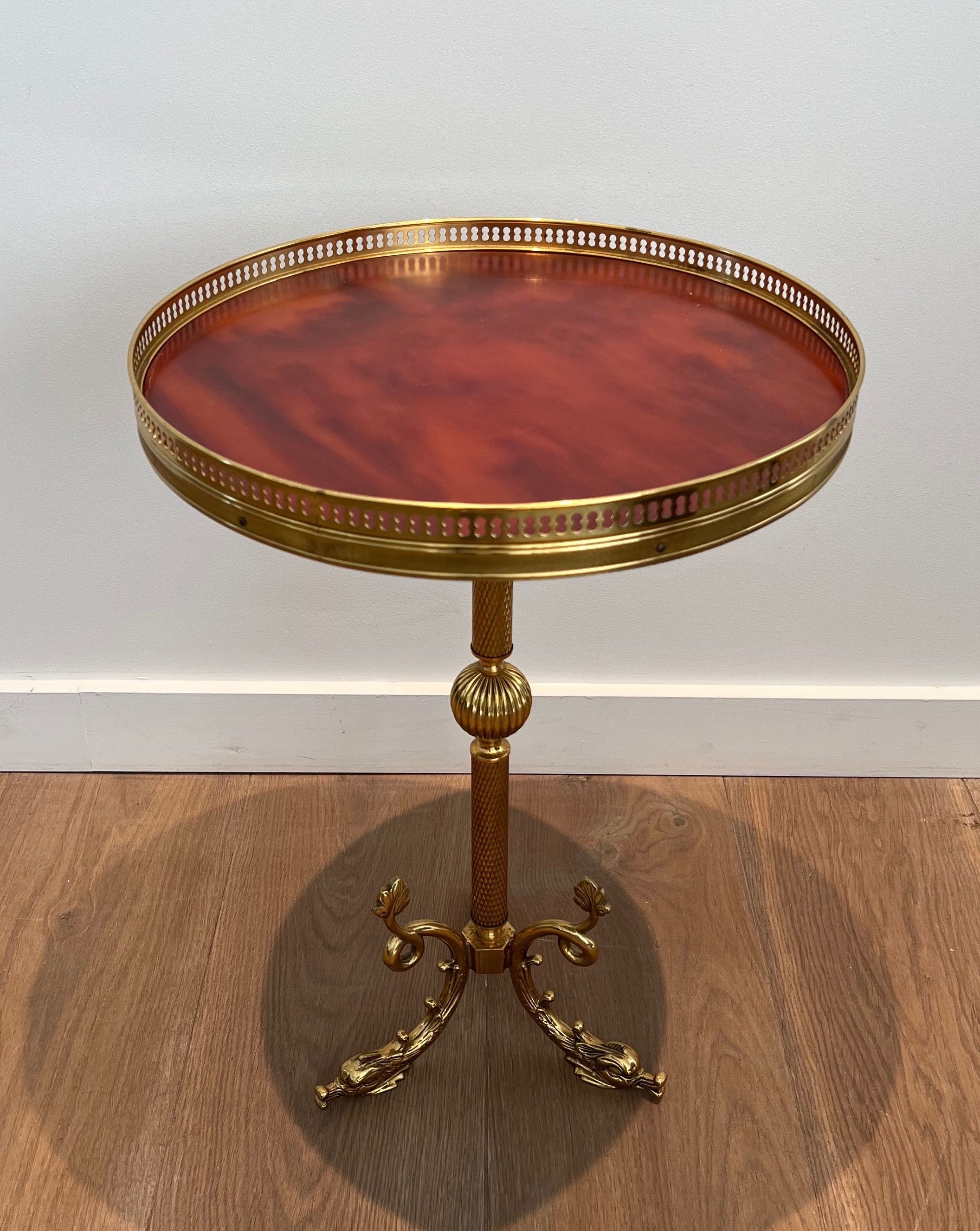 A round, neoclassical-style brass side table with a base decorated with dolphins and a red celluloid top encircled by an openwork gallery, attributed to the House of Jansen.