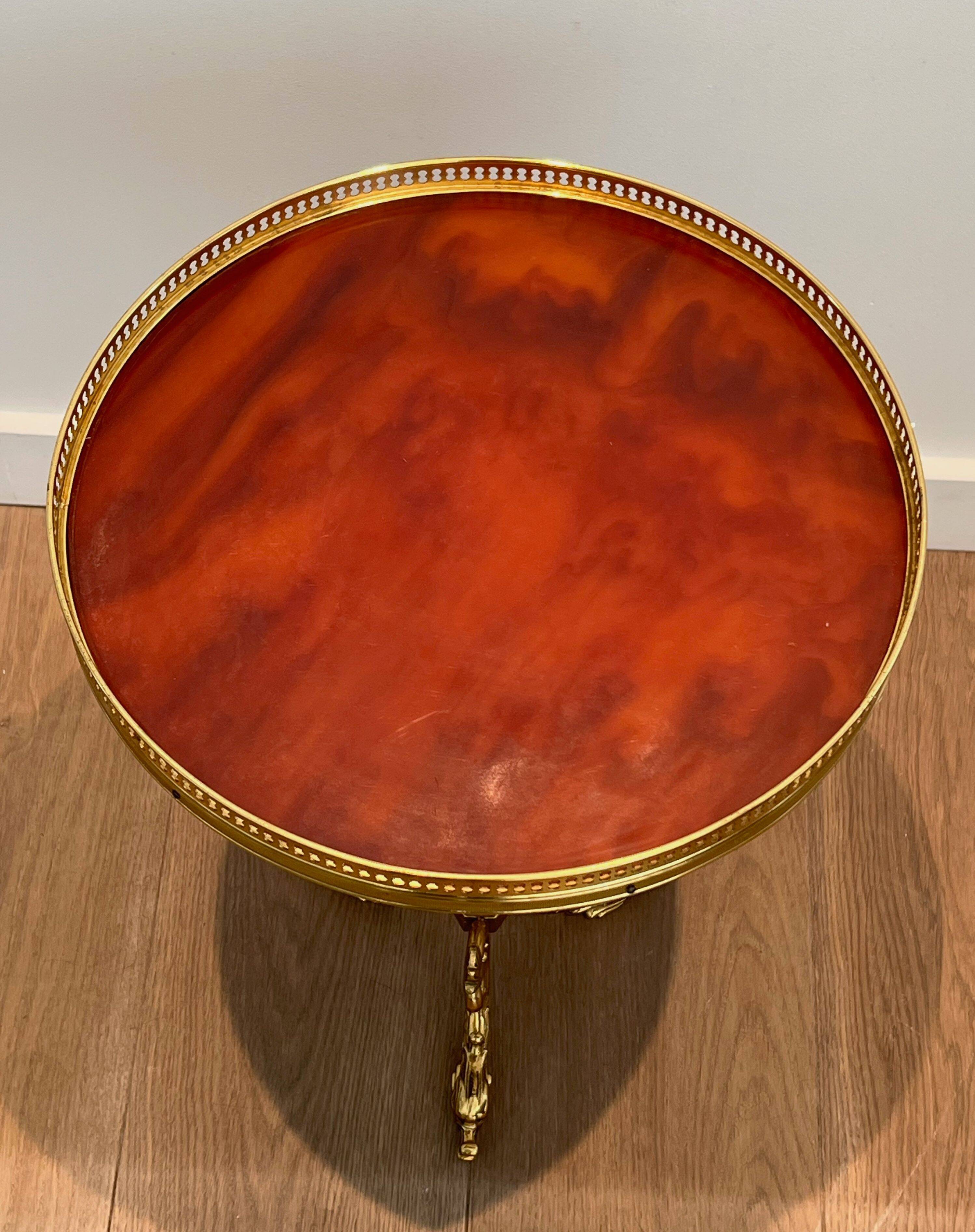 A round, neoclassical-style brass side table with a base decorated with dolphins and a red celluloid top encircled by an openwork gallery, attributed to the House of Jansen.