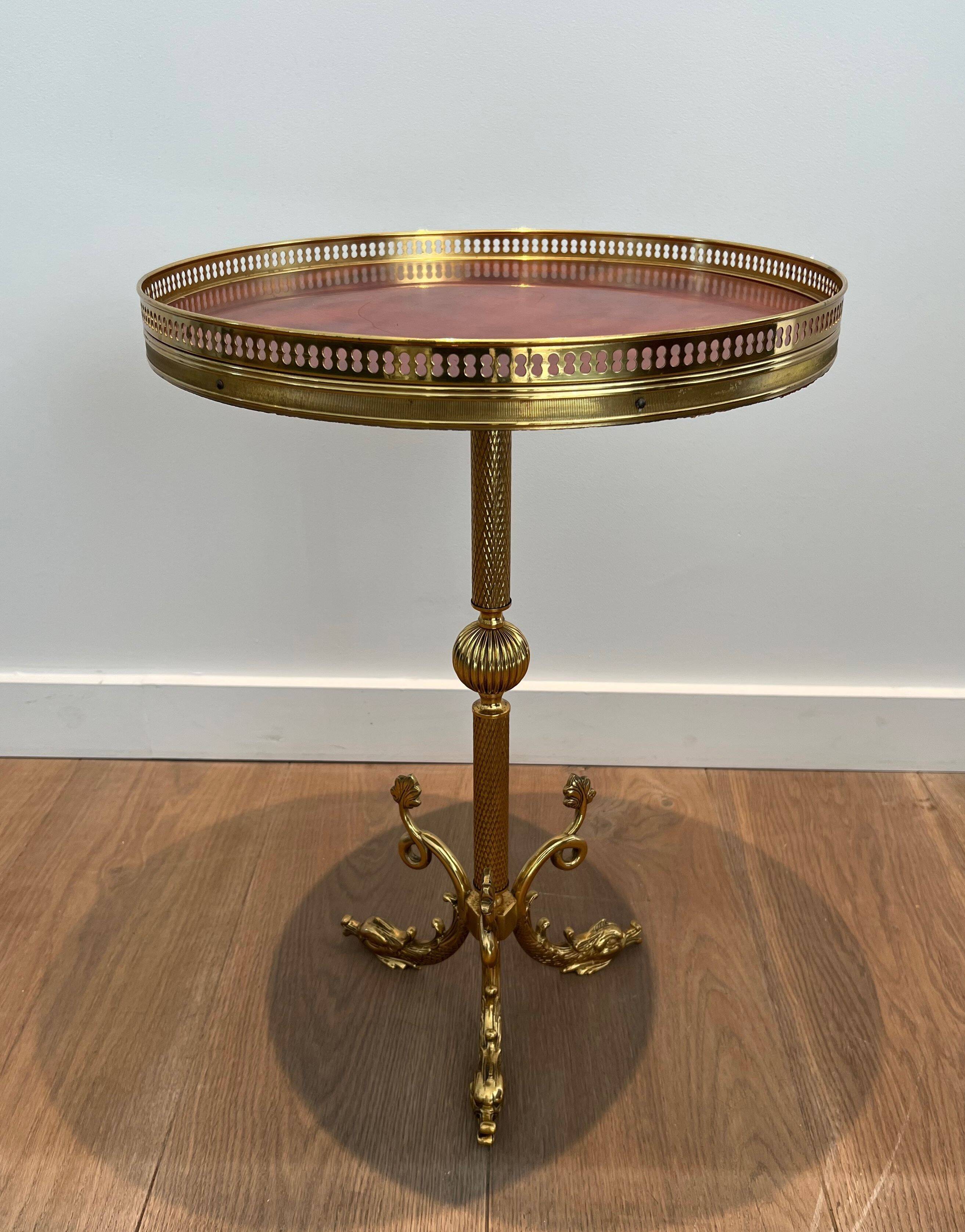 A round, neoclassical-style brass side table with a base decorated with dolphins and a red celluloid top encircled by an openwork gallery, attributed to the House of Jansen.