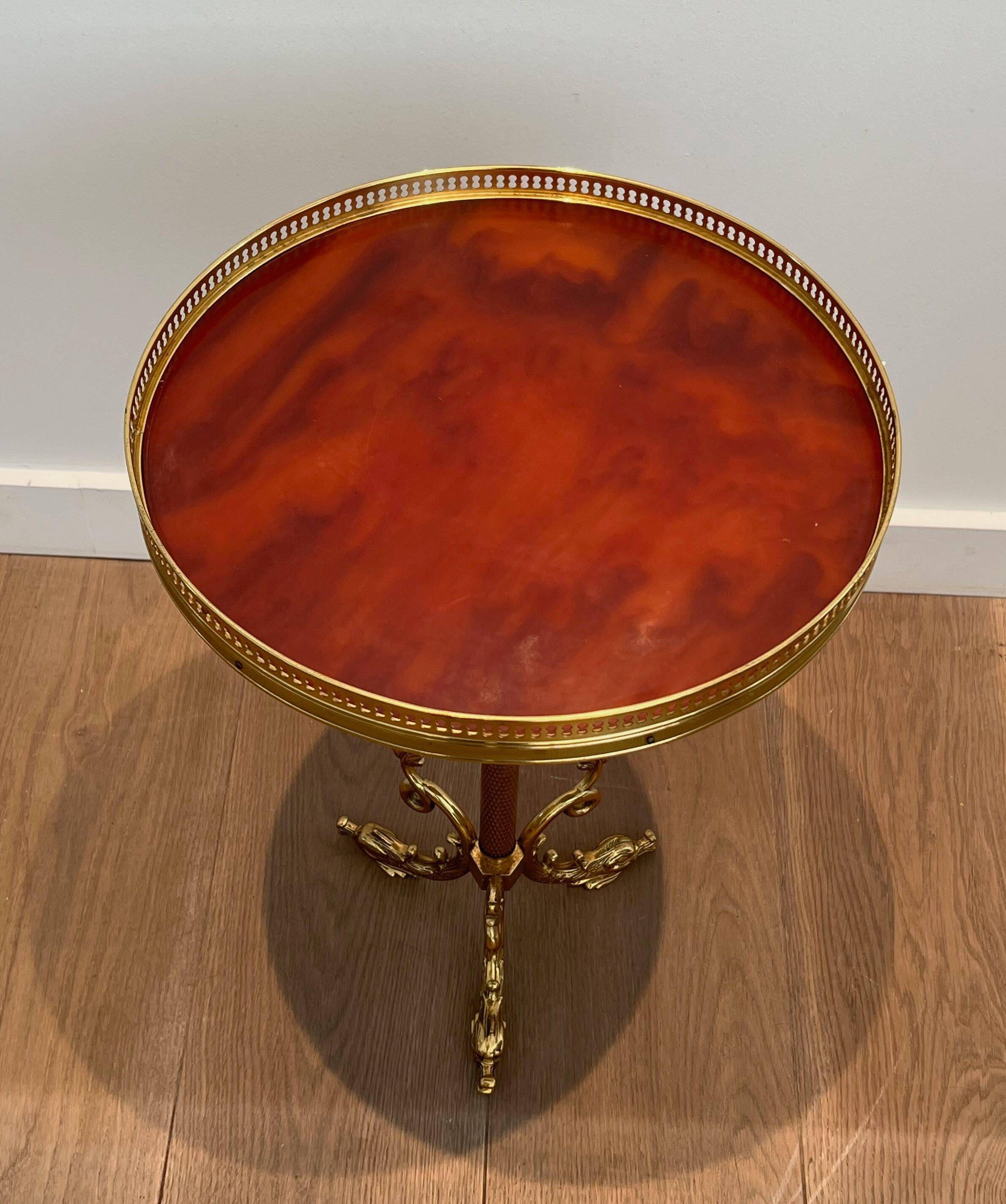 A round, neoclassical-style brass side table with a base decorated with dolphins and a red celluloid top encircled by an openwork gallery, attributed to the House of Jansen.