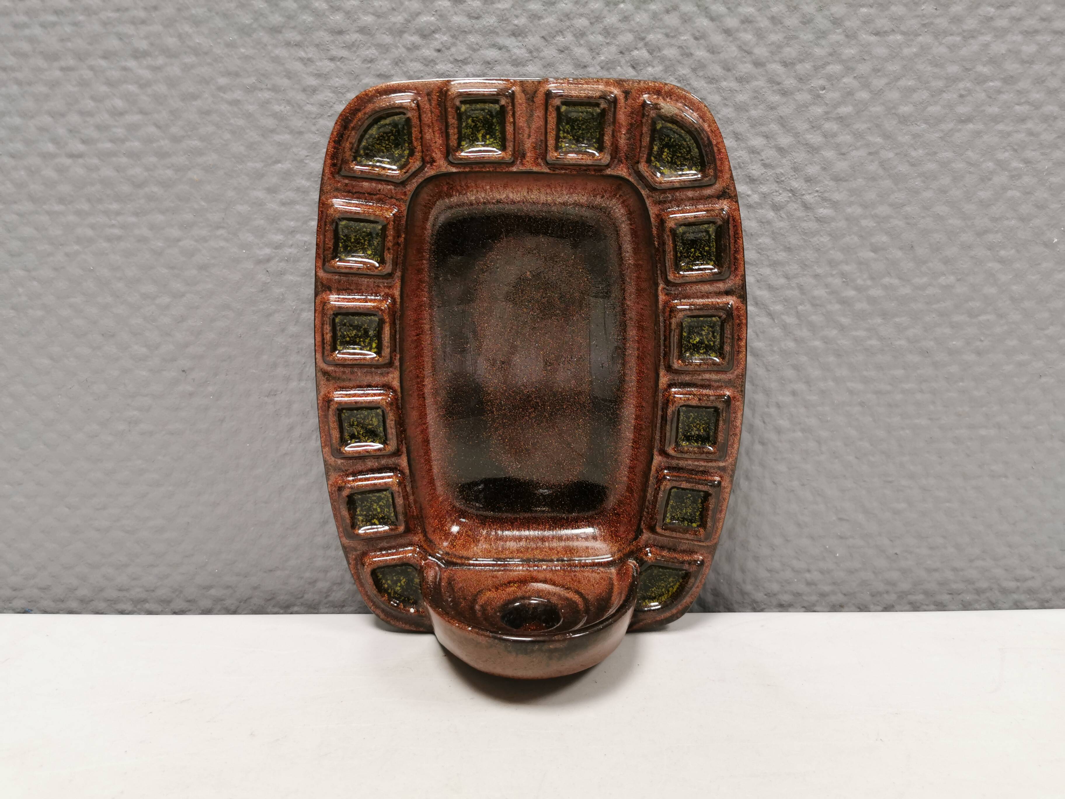 Wall-mounted candleholder in earthy, greenish, golden colours and shades.