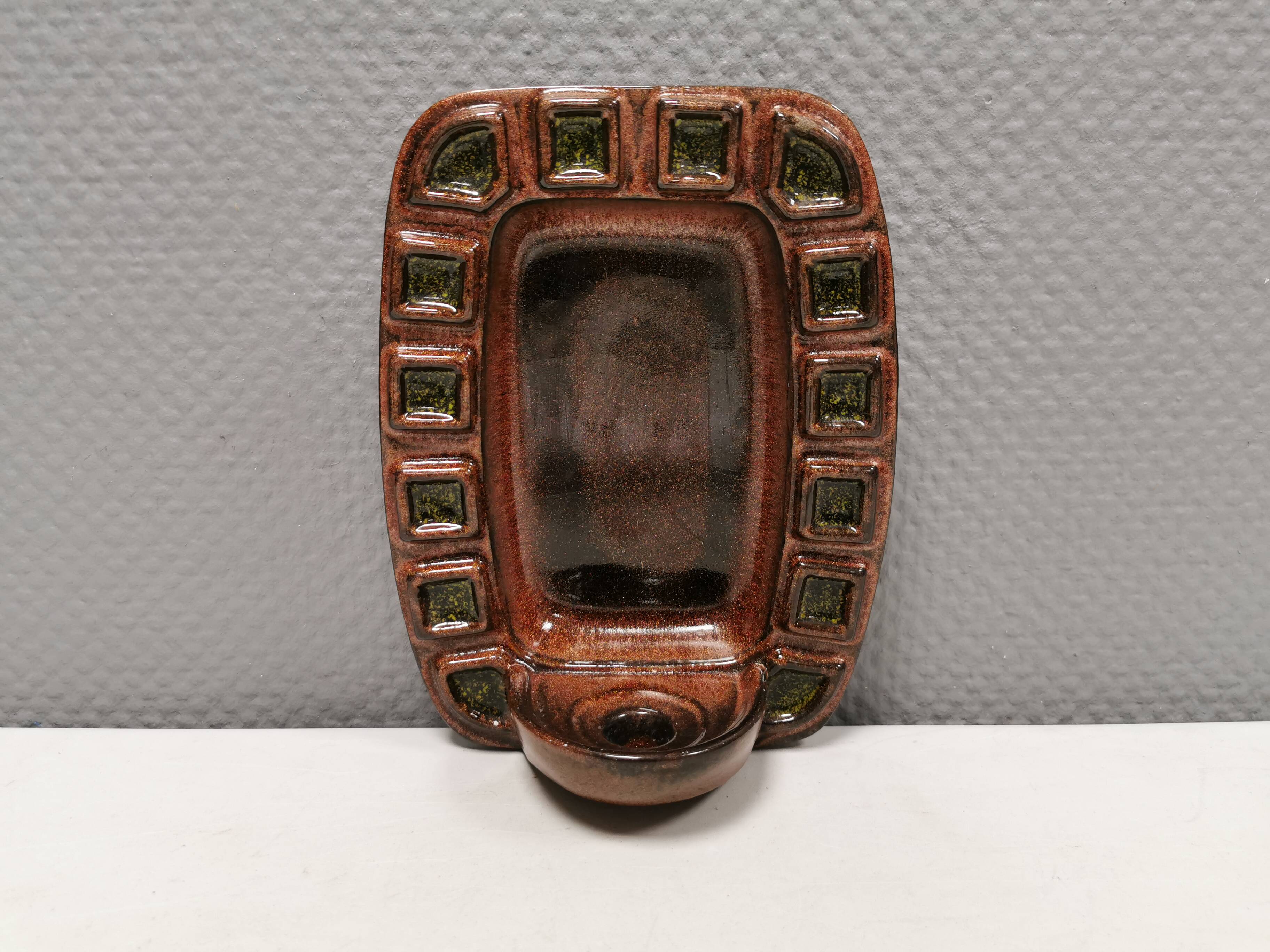 Wall-mounted candleholder in earthy, greenish, golden colours and shades.