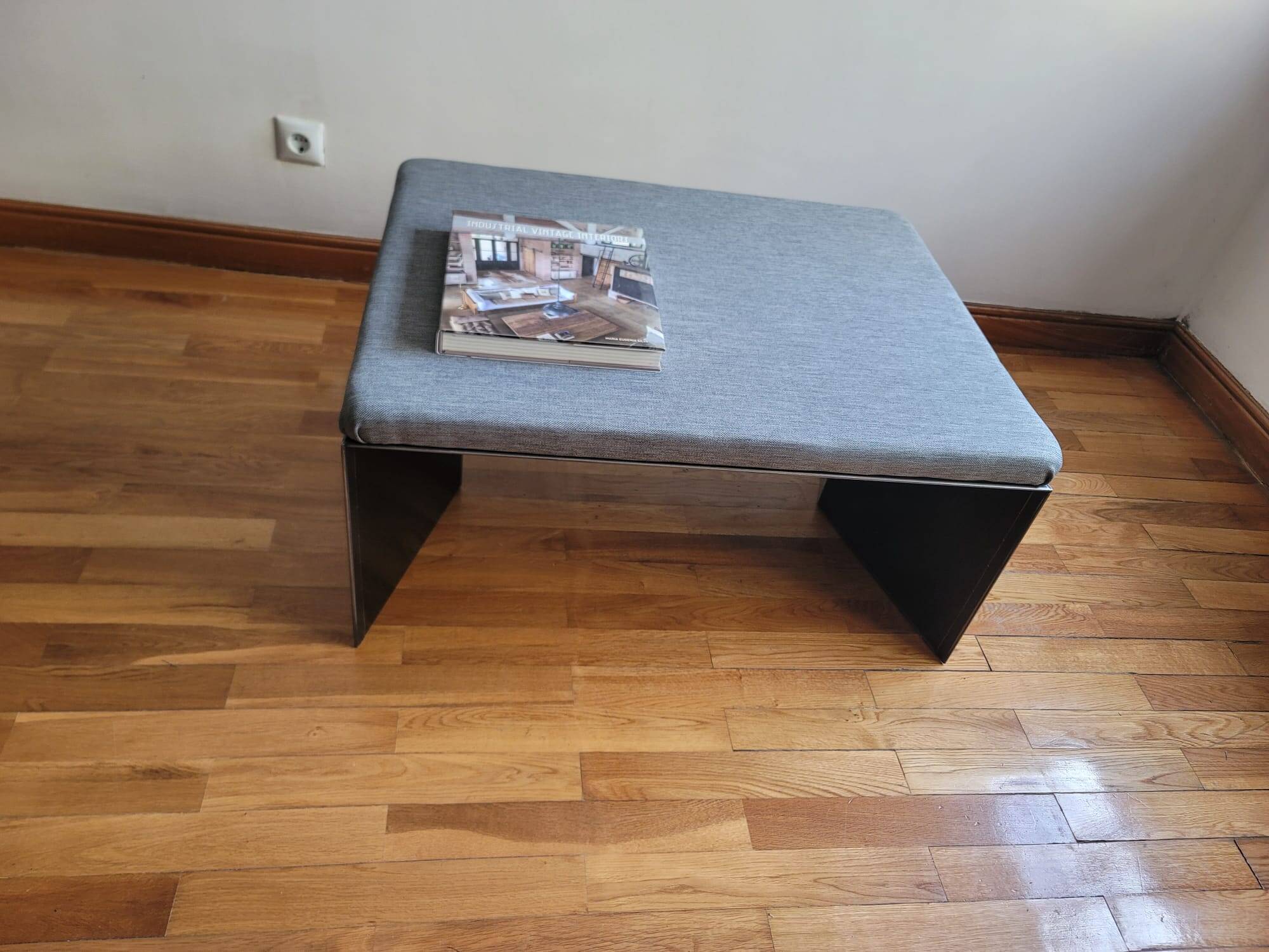 Minimalist/industrial coffee table with a black metal frame and a grey fabric top.