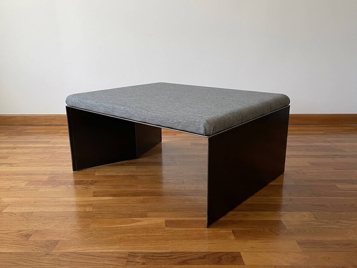 Minimalist/industrial coffee table with a black metal frame and a grey fabric top.