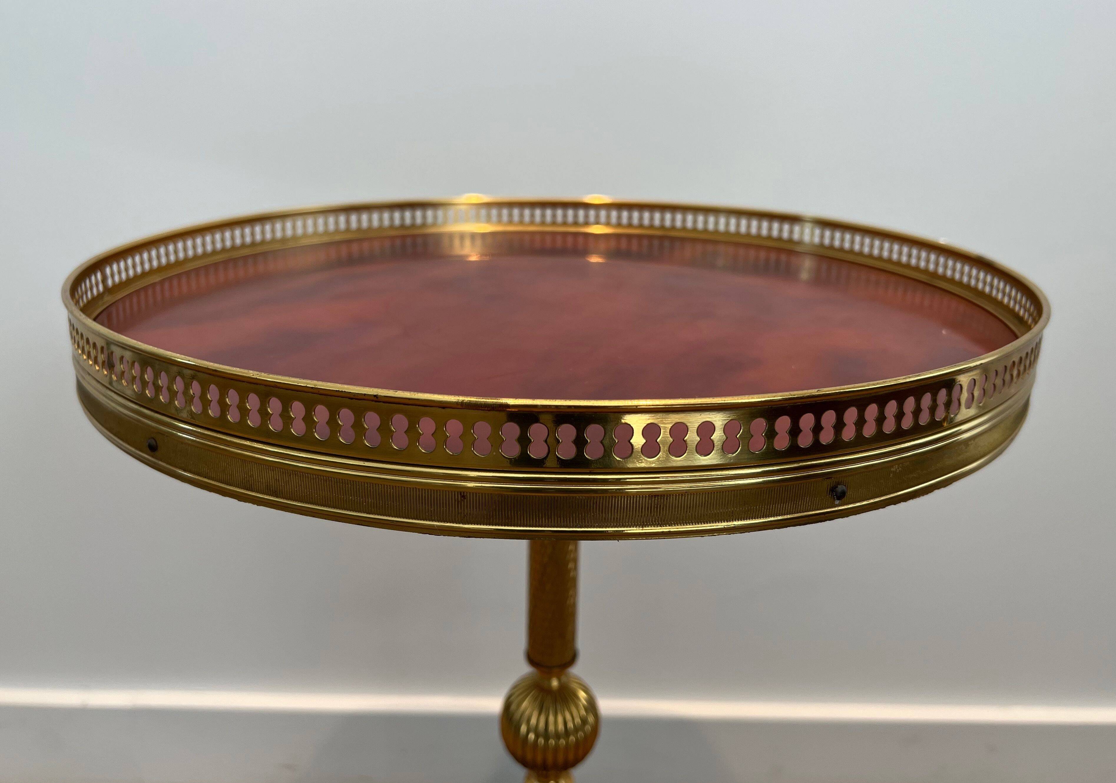 A round, neoclassical-style brass side table with a base decorated with dolphins and a red celluloid top encircled by an openwork gallery, attributed to the House of Jansen.