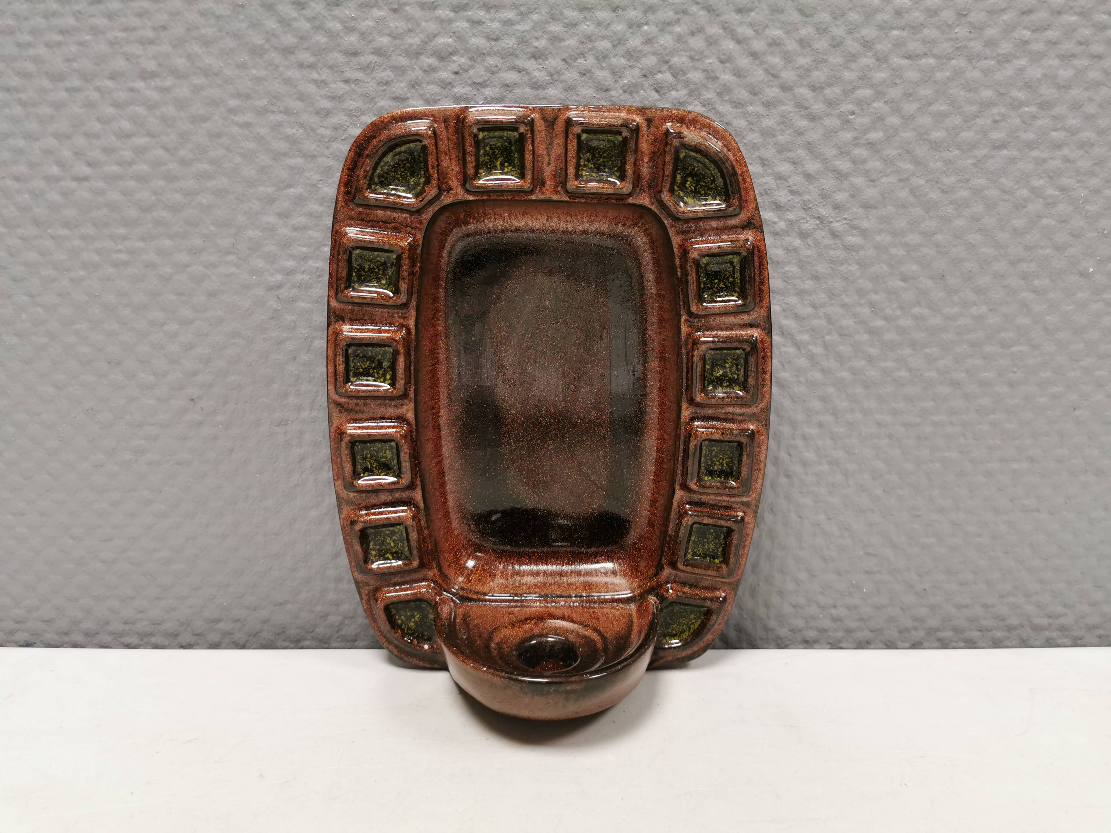 Wall-mounted candleholder in earthy, greenish, golden colours and shades.