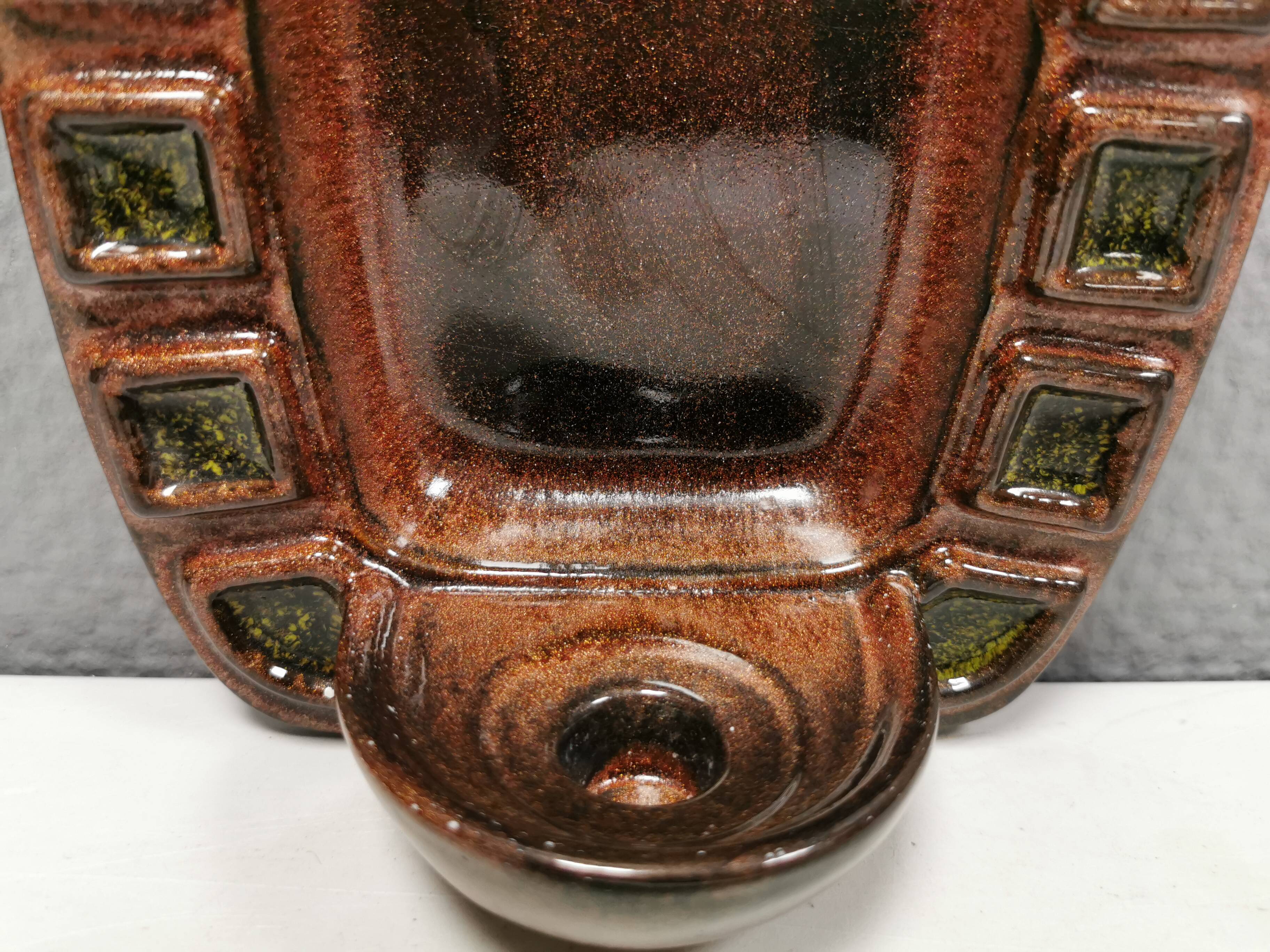 Wall-mounted candleholder in earthy, greenish, golden colours and shades.