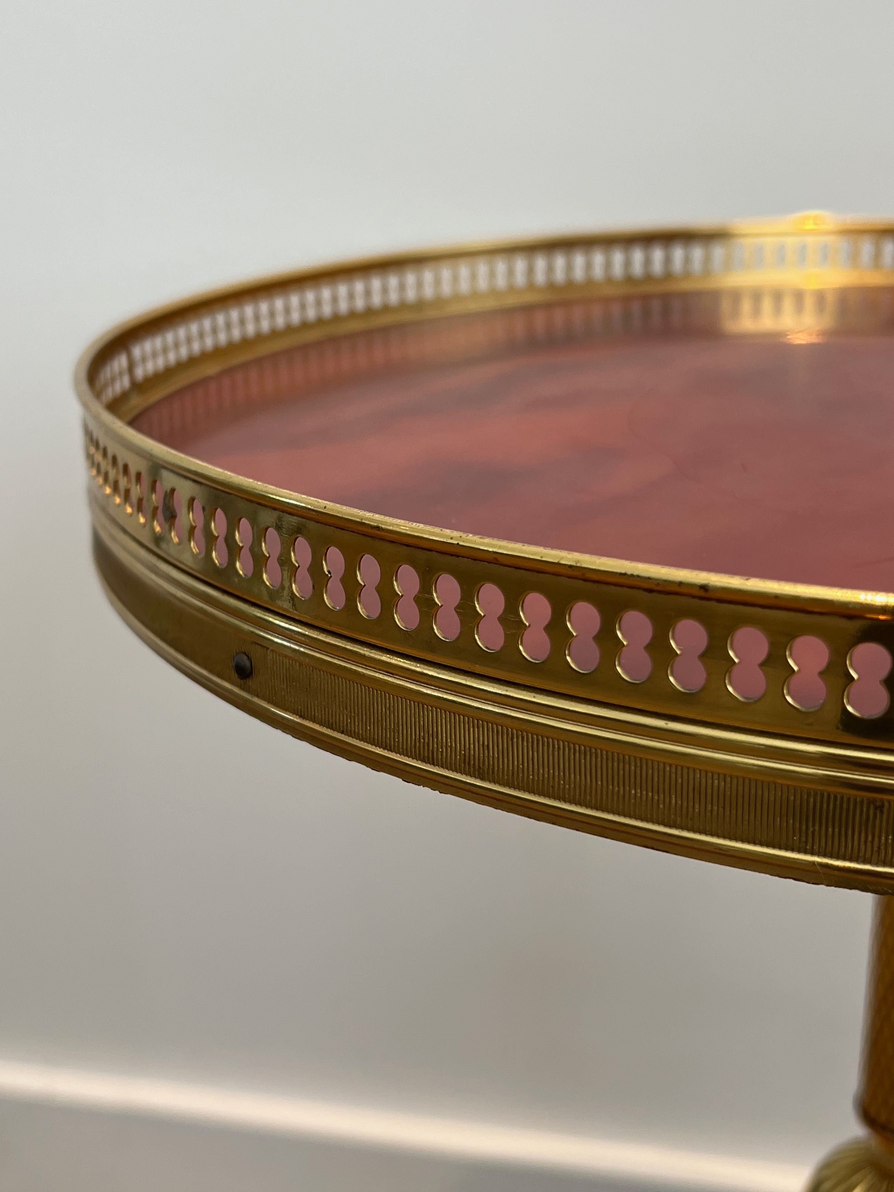 A round, neoclassical-style brass side table with a base decorated with dolphins and a red celluloid top encircled by an openwork gallery, attributed to the House of Jansen.