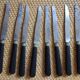 A set of 8 knives with black wooden handles and bolsters.