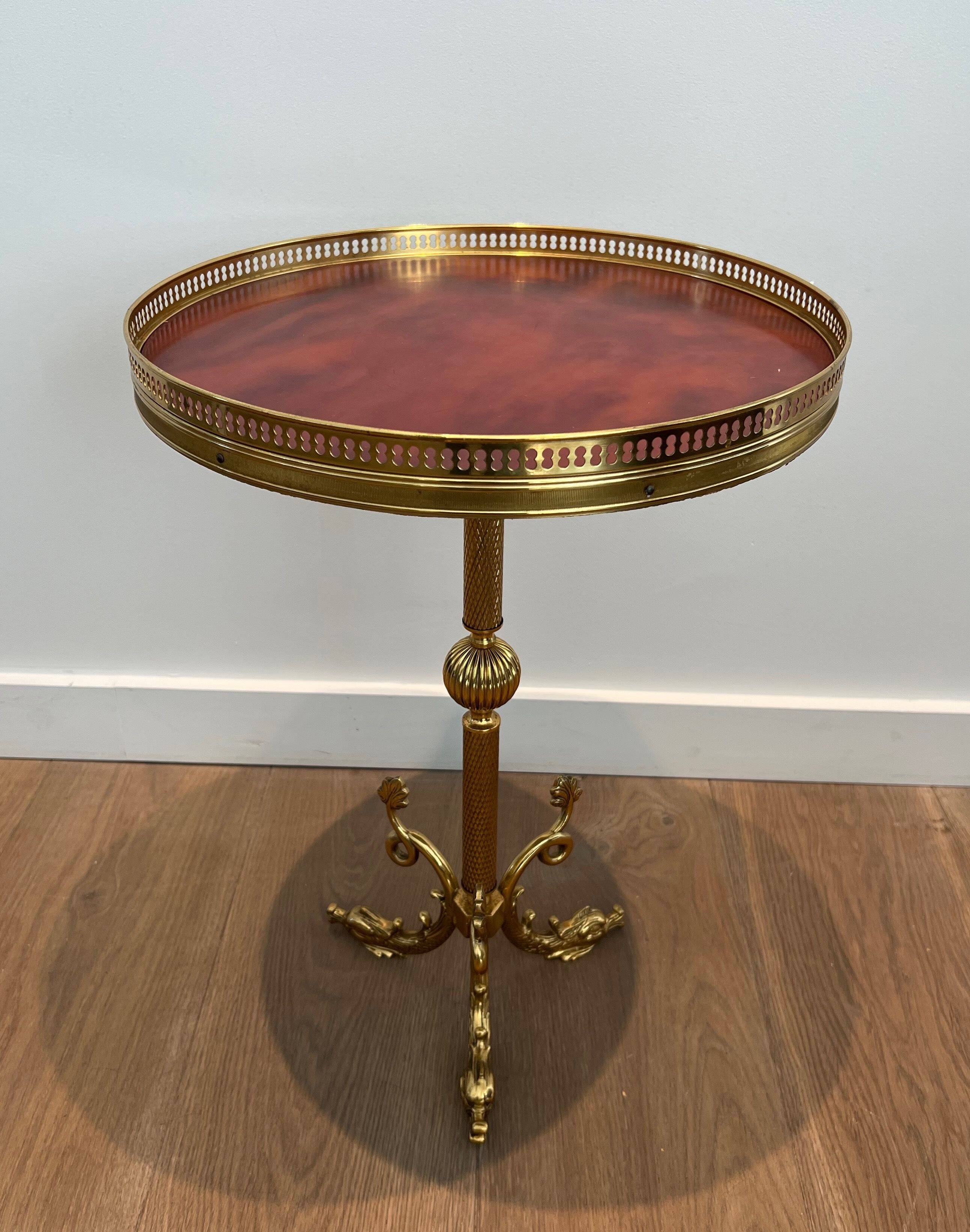 A round, neoclassical-style brass side table with a base decorated with dolphins and a red celluloid top encircled by an openwork gallery, attributed to the House of Jansen.