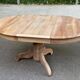 Round extendable dining table stripped in solid raw walnut with a central leg.
