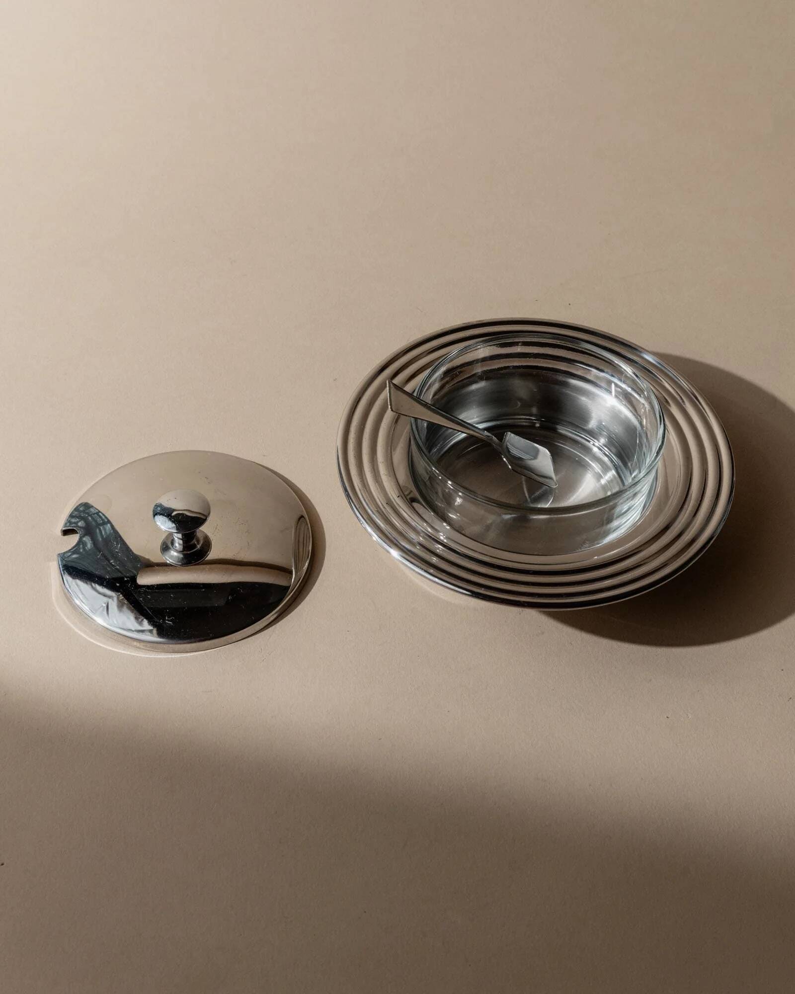 Vintage 1970s condiment pot made of glass and stainless steel with a lid, complete with a matching spoon.