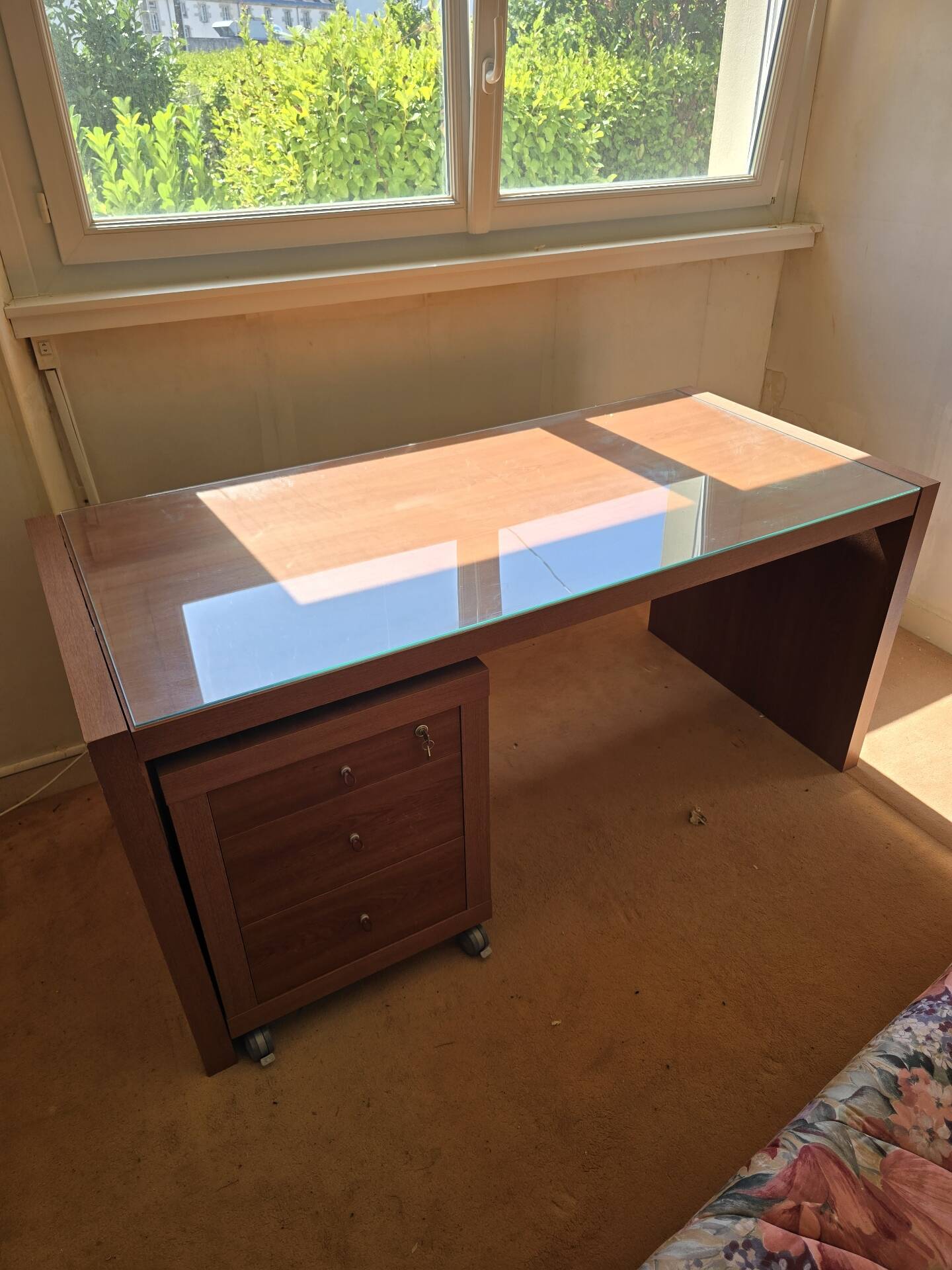 Large wooden desk with a glass top and a wheeled drawer unit.