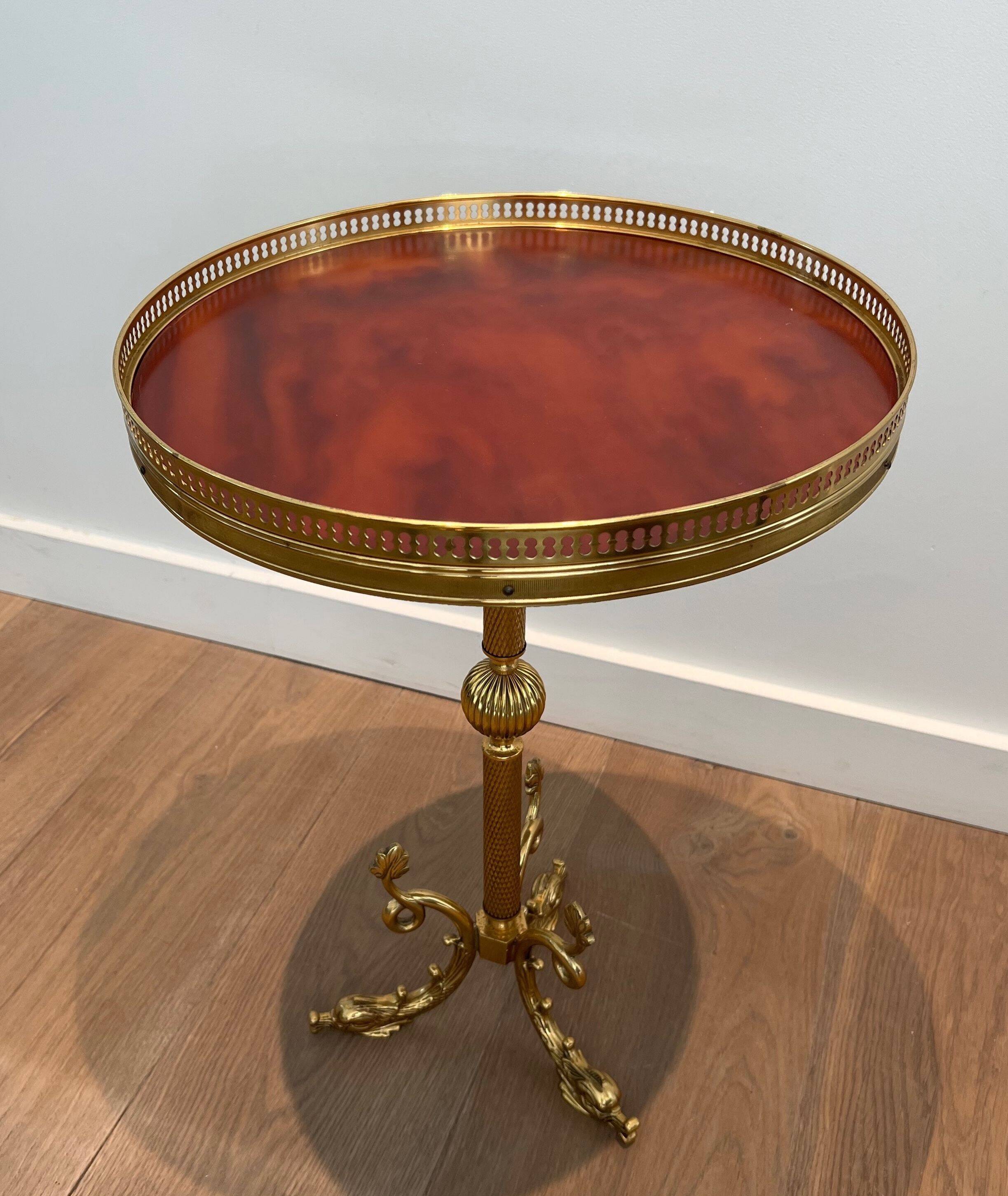 A round, neoclassical-style brass side table with a base decorated with dolphins and a red celluloid top encircled by an openwork gallery, attributed to the House of Jansen.